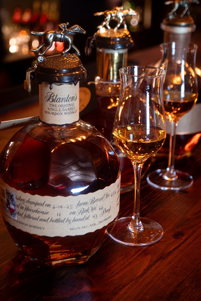 Bottle of Blanton's Single Barrel Bourbon Whiskey with its iconic horse-and-jockey stopper on a wooden bar at Highlands Lounge, with a tulip-shaped tasting glass beside it